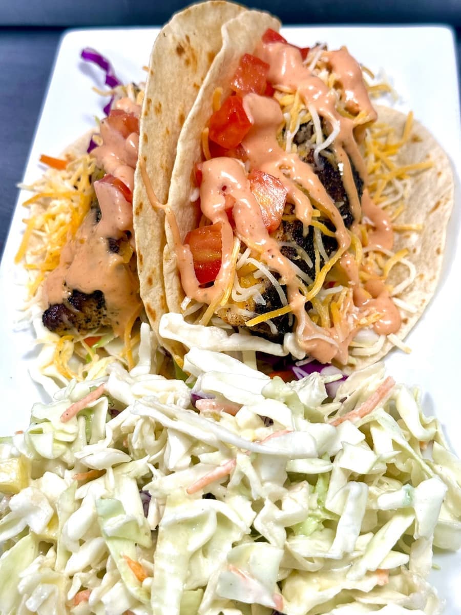Two blackened fish tacos in flour tortillas with shredded cabbage, cheese, diced tomato and an aioli drizzle, next to a side of coleslaw on a white plate